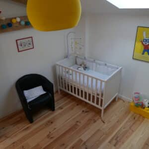 Chambre bébé lumineuse avec parquet bois clair, berceau blanc, fauteuil noir, suspension jaune et affiche lapin super-héros.