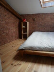 Chambre mansardée lumineuse avec mur en briques rouges, plancher en bois clair, lit simple en bois et étagère, fenêtre de toit.