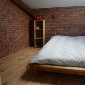 Chambre mansardée lumineuse avec mur en briques rouges, plancher en bois clair, lit simple en bois et étagère, fenêtre de toit.