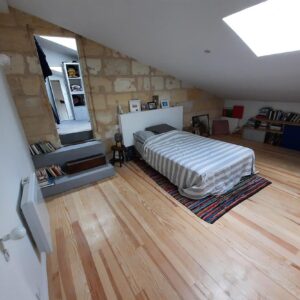 Chambre mansardée lumineuse avec mur en pierre apparente, lit rayé, sol en bois clair. Escalier avec livres vers dressing.