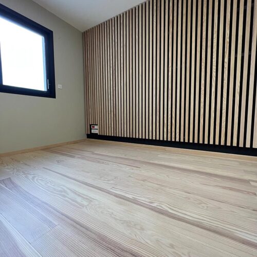 Chambre moderne vide avec parquet clair et mur d'accent en lattes de bois verticales foncées. Fenêtre noire à gauche.