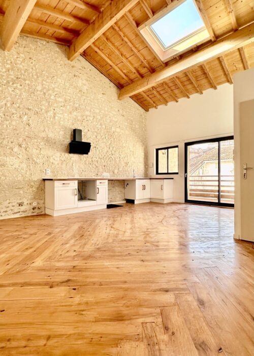 Loft rénové avec mur en pierres apparentes, plafond et sol en bois clair. Cuisine nue et grande baie vitrée offrant vue sur toits.