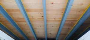 Plafond moderne en bois clair avec poutres de soutien métalliques bleu-gris. Les planches apparentes offrent une texture chaleureuse.