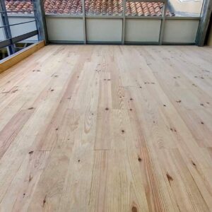 Plancher en bois clair brut posé sur une mezzanine, avec balustrade métallique et vue sur des toits en tuiles. Espace en rénovation.