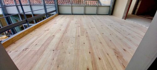 Plancher bois clair neuf sur mezzanine, vue sur tuiles de toit et balustrade métallique. Espace intérieur en rénovation.