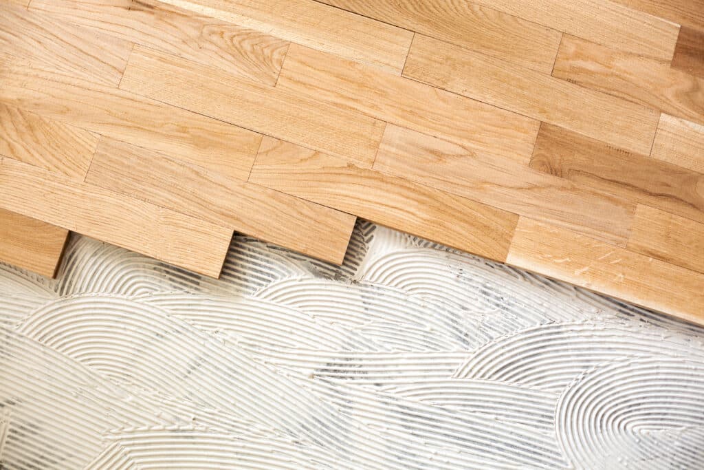 Planches de parquet en bois clair sur adhésif blanc ondulé, installation de revêtement de sol en cours.