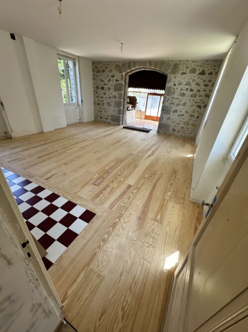 Pièce rénovée avec parquet en pin clair, mur en pierres brutes et carreaux damier. Arche menant à une véranda.