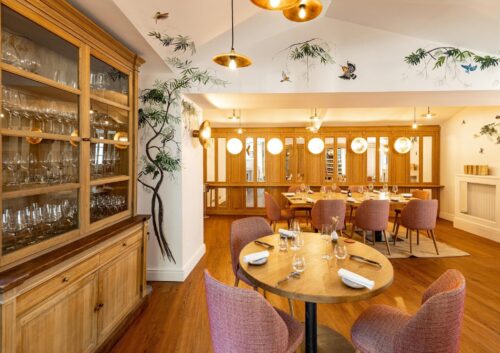 Élégante salle à manger de restaurant. Muraux d'oiseaux et branches, grand buffet en bois, tables rondes avec chaises roses, lumière dorée.