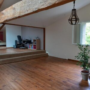 Salon lumineux avec mezzanine bureau, parquet bois, poutres apparentes, fenêtre ouverte et plante verte. Ambiance rustique chic.
