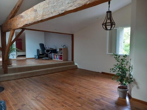 Salon lumineux avec mezzanine bureau, parquet bois, poutres apparentes, fenêtre ouverte et plante verte. Ambiance rustique chic.