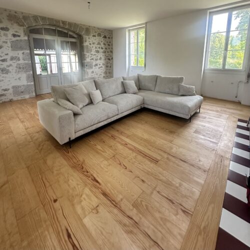 Salon moderne avec grand canapé d'angle gris clair, parquet en bois blond, mur de pierres et baies vitrées lumineuses.