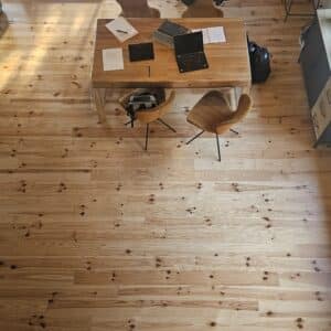 Vue plongeante d'un salon avec parquet en pin des landes, canapé, table de travail ouverte avec ordinateur portable, et grandes fenêtres donnant sur l'extérieur.
